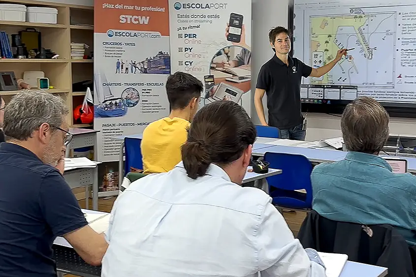Un instructor de Escola Port señala una carta náutica en la pizarra durante una clase presencial del curso de Patrón de yate, con alumnos tomando apuntes en el aula del Port Olímpic.