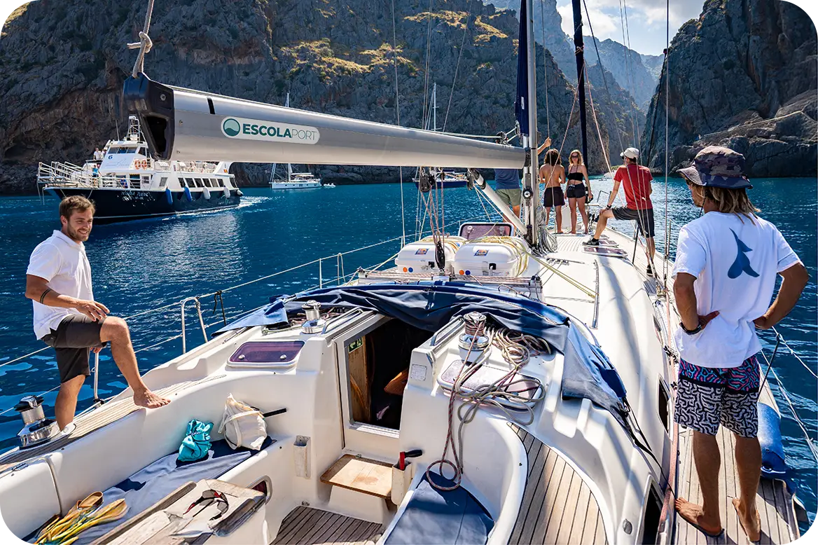 Alumnos de Escola Port realizando las prácticas del curso PER a bordo en un velero en Cala Macarella (Menorca), con el agua turquesa y los acantilados de fondo.