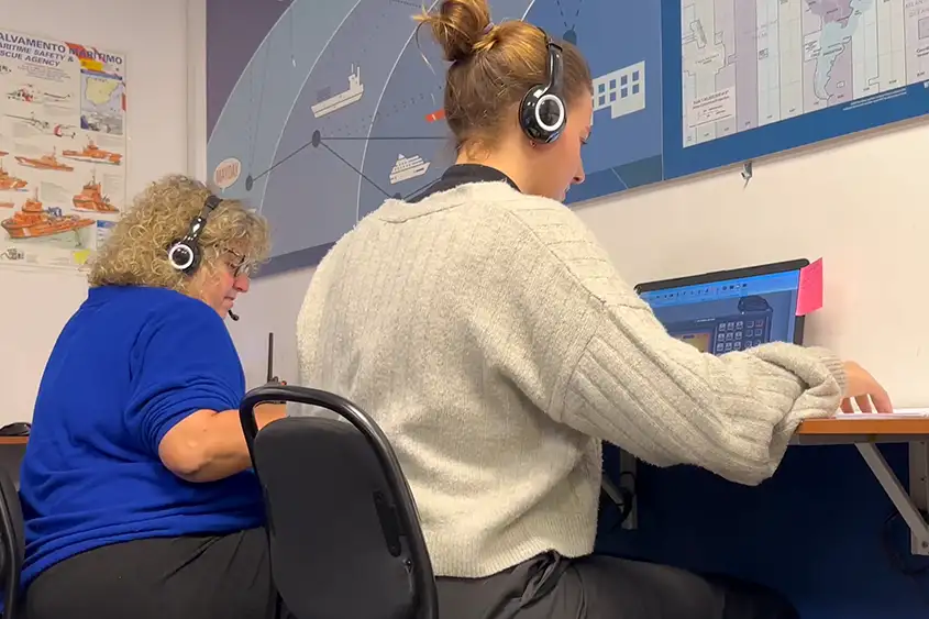 Dos alumnas con auriculares practican una comunicación de radio en el simulador, con el mapa de zonas del SMSSM en la pared.